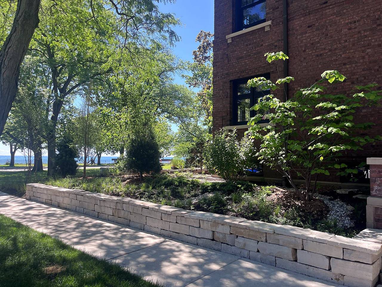 A native garden behind a dry laid limestone wall beside a brick lakefront house, with Lake Michigan through the trees